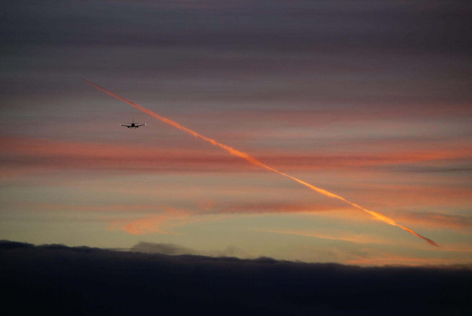 The silhouette of a jet airliner seen head-on against a sunset sky, its contrail glowing orange, the kind of sustained high-altitude track a real airborne node leaves behind.