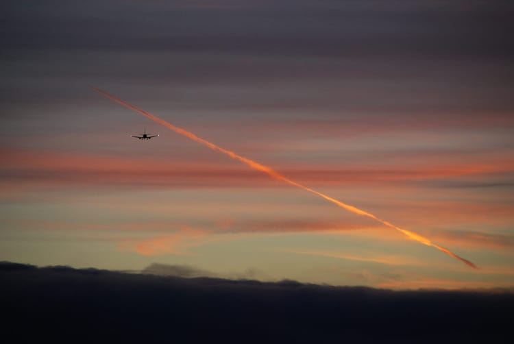 The silhouette of a jet airliner seen head-on against a sunset sky, with its contrail glowing orange in the last light.