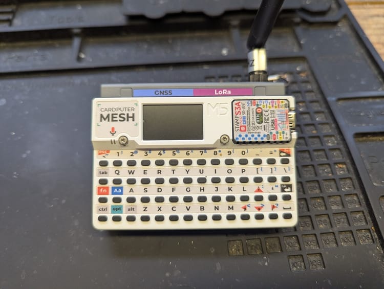 M5Stack Cardputer with LoRa and GNSS radio module clipped onto the top edge, showing the full QWERTY keyboard and screen