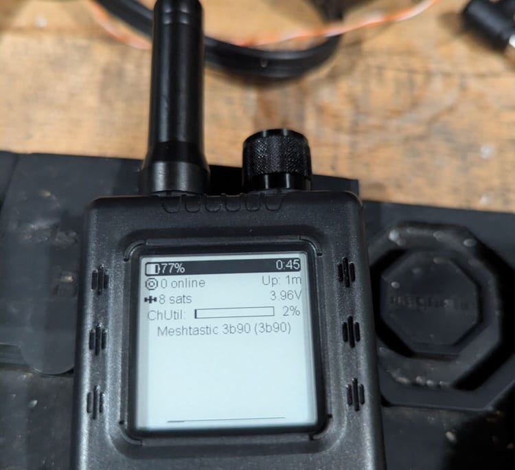 A ThinkNode Meshtastic handheld node on a workbench, its screen showing 77% battery, 8 GPS satellites, 2% channel utilization, and the node name Meshtastic 3b90