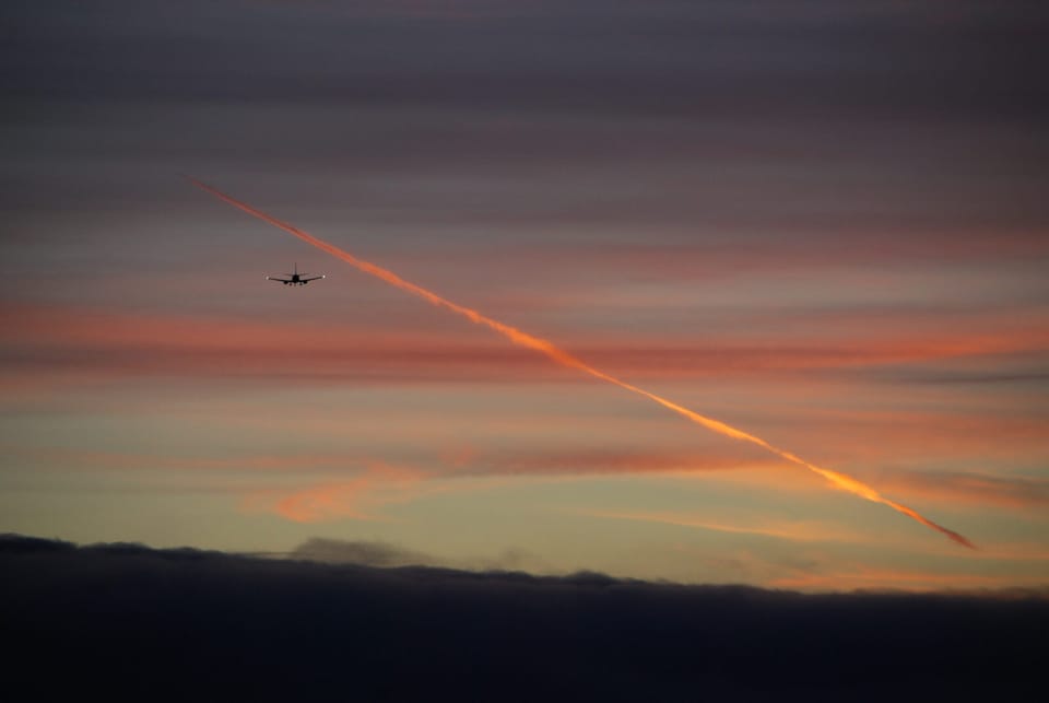 The silhouette of a jet airliner seen head-on against a sunset sky, with its contrail glowing orange in the last light.