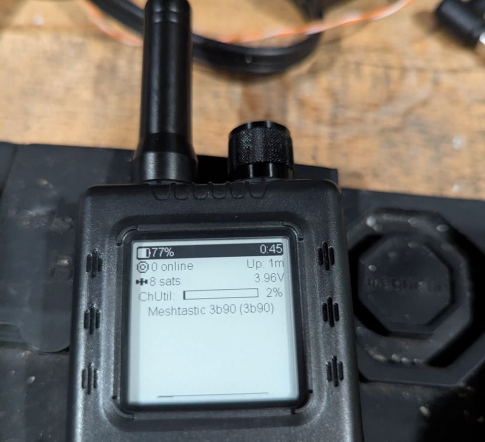 A ThinkNode Meshtastic handheld node on a workbench, its screen showing 77% battery, 8 GPS satellites, 2% channel utilization, and the node name Meshtastic 3b90
